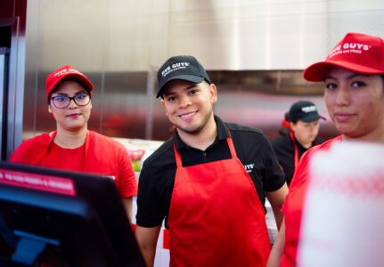 Three Five Guys crew members smile while posing for a photograph inside the kitchen.