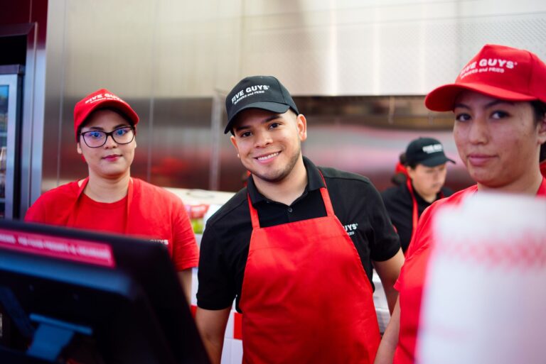 Three Five Guys crew members smile while posing for a photograph inside the kitchen.